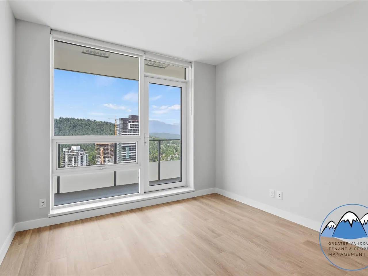 Bedroom with a sliding balcony door opening to forest and city views, with pale hardwood flooring.